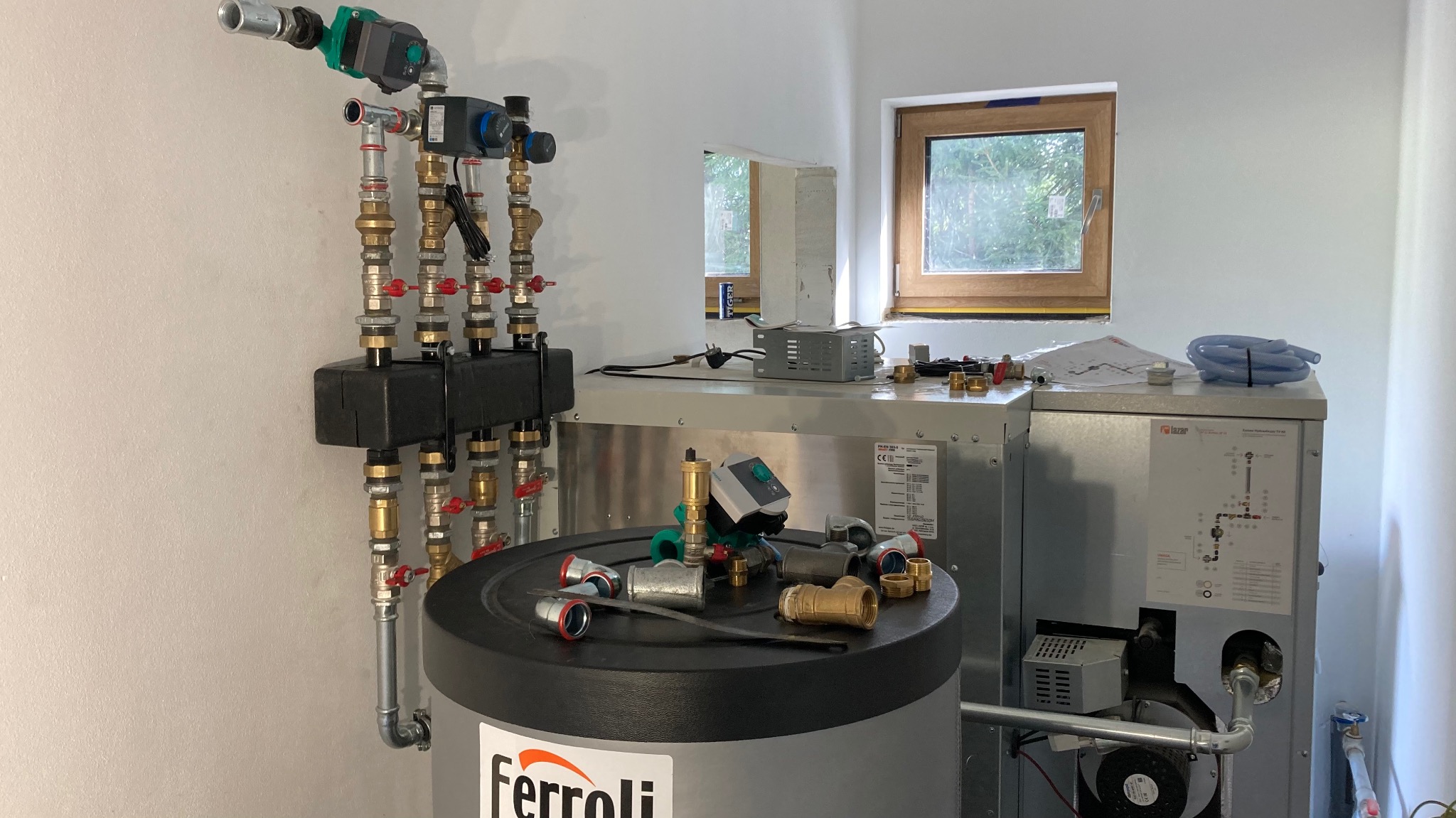 Nowa instalacja grzewcza w kotłowni, widoczne rury, zawory, pompy i zbiornik Ferroli z akcesoriami na pokrywie, małe okno w tle.