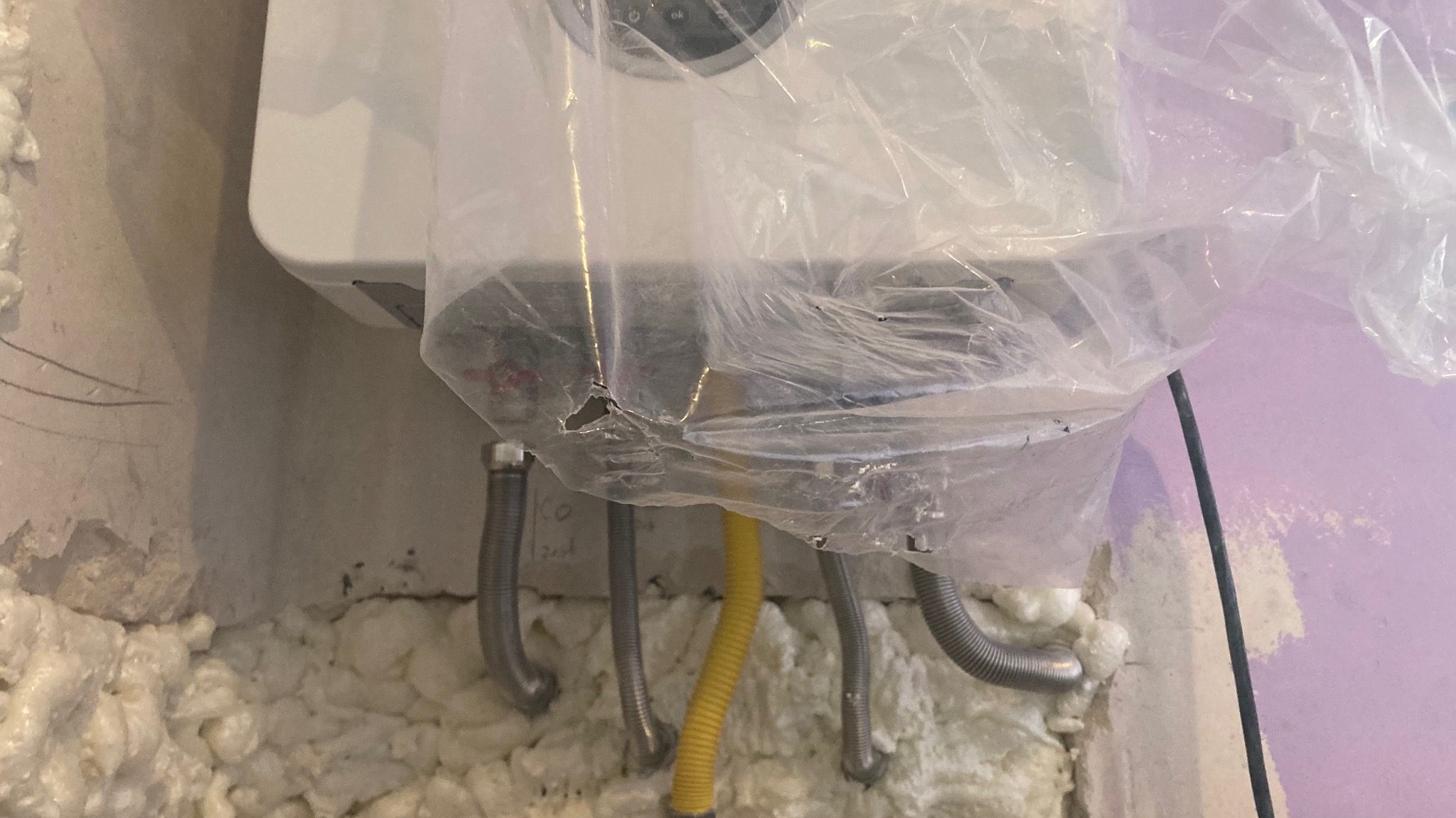 Nowy, biały piec gazowy, zabezpieczony folią ochronną, z podłączonymi elastycznymi przewodami gazowymi i instalacją elektryczną, zamontowany na ścianie z widoczną pianką montażową.