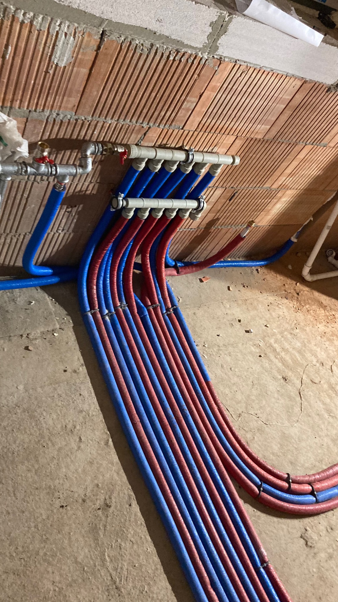 Rozdzielacz instalacji wodnej z rurami PEX w kolorze niebieskim i czerwonym, mocowany do ściany z cegły dziurawki, widoczne elementy montażowe i fragment instalacji z zaworem.