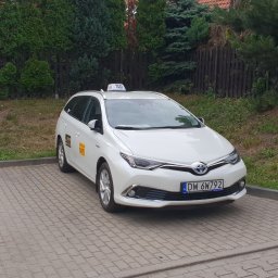 GO CAR TAXI MAŁGORZATA MAJCHROWICZ - Biała Toyota Auris kombi z oznaczeniami taxi, zaparkowana na brukowanym placu, z widocznym numerem bocznym 1475 i tablicą rejestracyjną DW 6W792, na tle drewnianego płotu i zieleni.