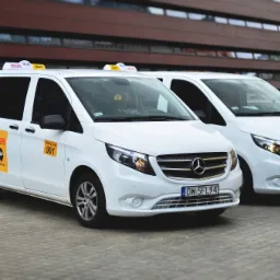 Dwa białe busy Mercedes-Benz Vito z oznaczeniami taxi Wicar, zaparkowane przed nowoczesnym budynkiem z ciemnego drewna i szkła we Wrocławiu, z widocznymi tablicami rejestracyjnymi DW 5FL94 i DW 1NM13.