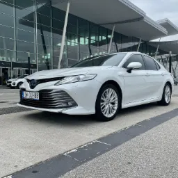 Biała Toyota Camry Hybrid zaparkowana przed nowoczesnym budynkiem lotniska z przeszkloną fasadą i charakterystycznymi daszkami.