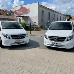 GO CAR TAXI MAŁGORZATA MAJCHROWICZ - Dwa białe busy Mercedes Vito zaparkowane na brukowanym placu przed budynkiem z szyldem 'bajka artykuły dla dzieci'.