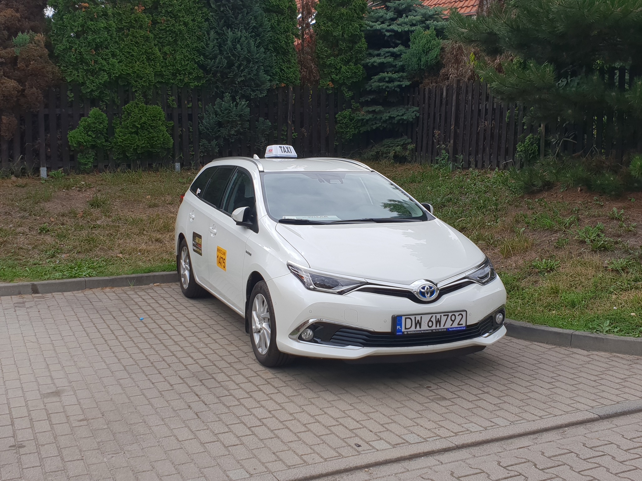 Biała Toyota Auris kombi z oznaczeniami taxi, zaparkowana na brukowanym placu, z widocznym numerem bocznym 1475 i tablicą rejestracyjną DW 6W792, na tle drewnianego płotu i zieleni.