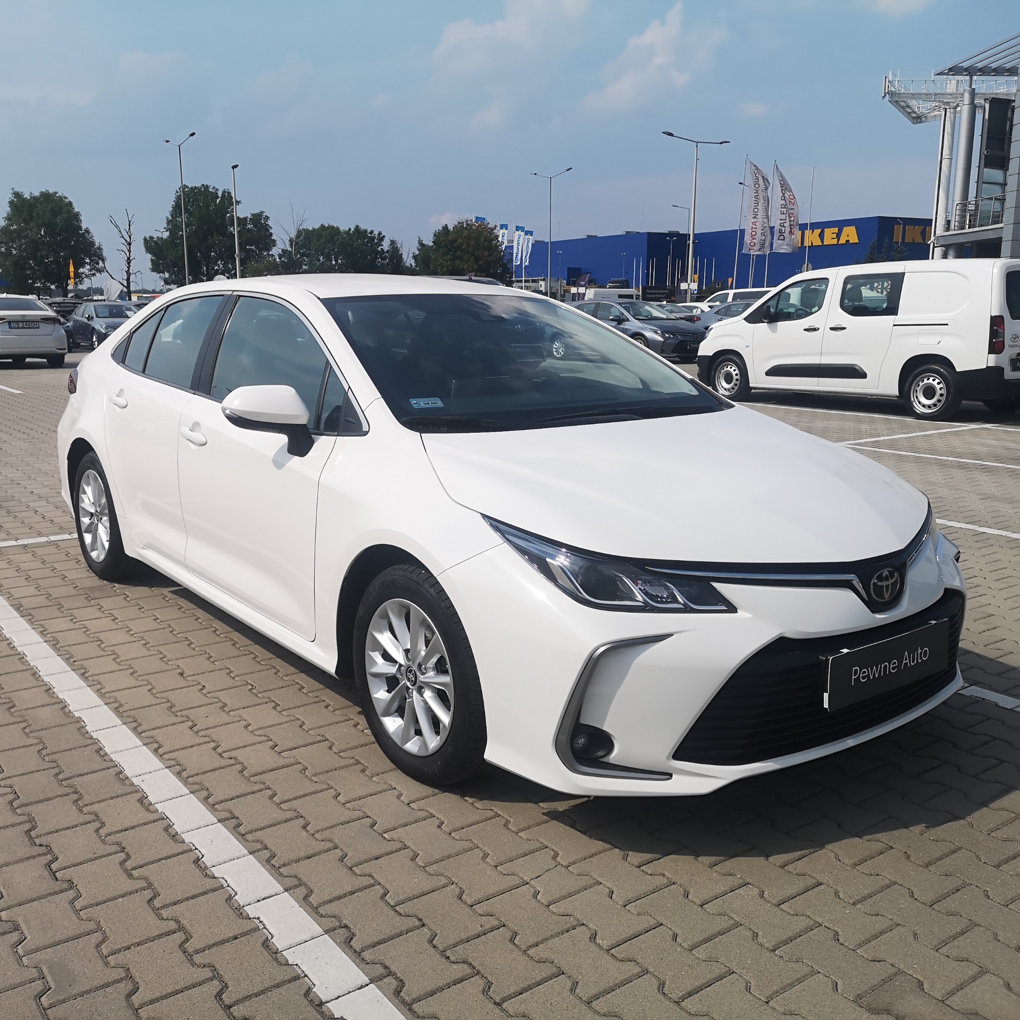 Biała Toyota Corolla zaparkowana na parkingu przed sklepem IKEA i Castorama, widoczny napis 'Pewne Auto' na tabliczce dealera.
