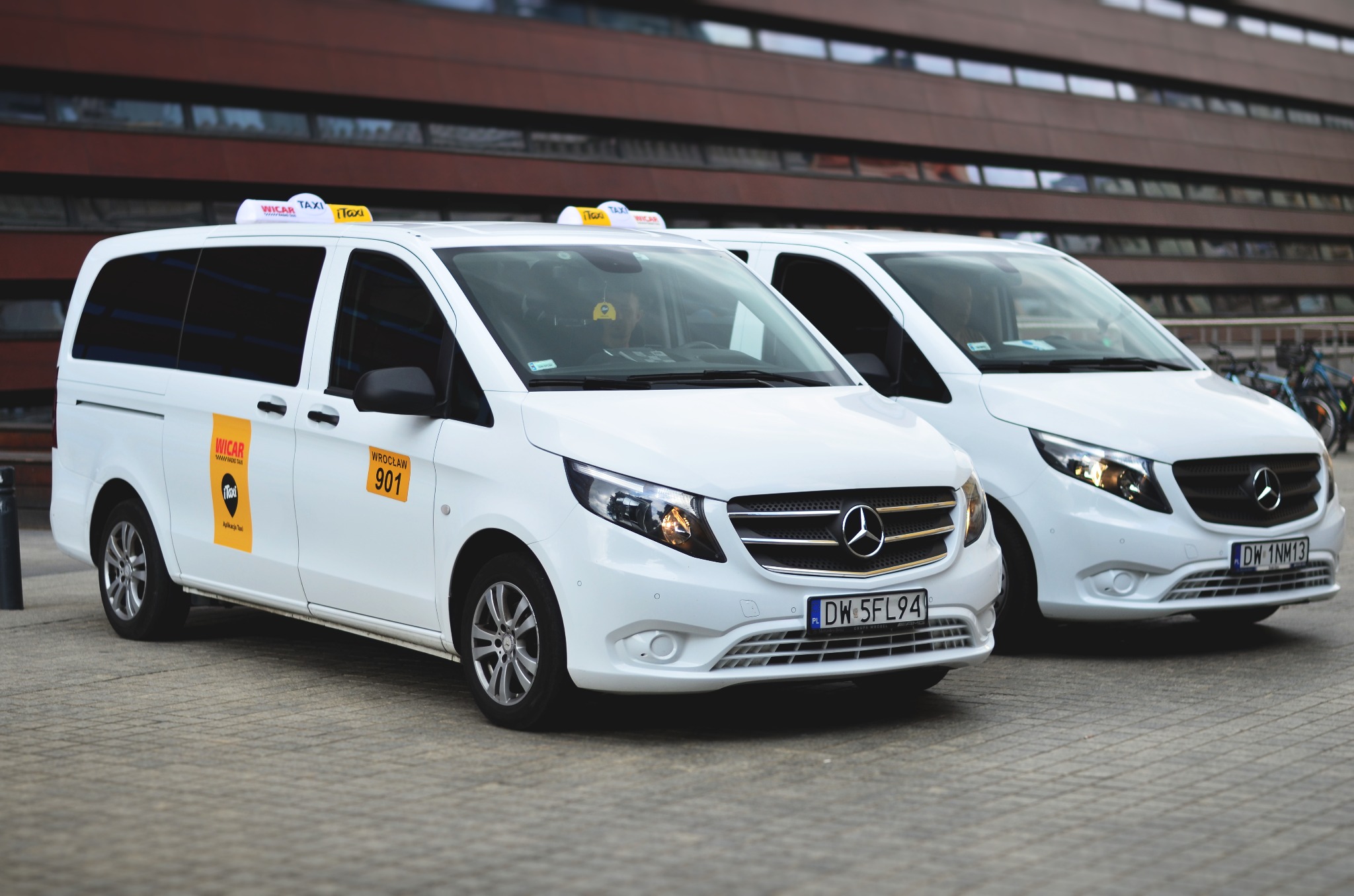 Dwa białe busy Mercedes-Benz Vito z oznaczeniami taxi Wicar, zaparkowane przed nowoczesnym budynkiem z ciemnego drewna i szkła we Wrocławiu, z widocznymi tablicami rejestracyjnymi DW 5FL94 i DW 1NM13.