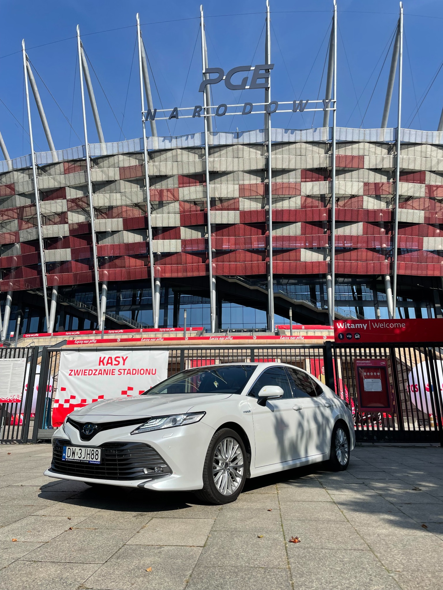 Biała Toyota Camry zaparkowana przed Stadionem Narodowym w Warszawie, w tle widoczny napis KASY ZWIEDZANIE STADIONU.