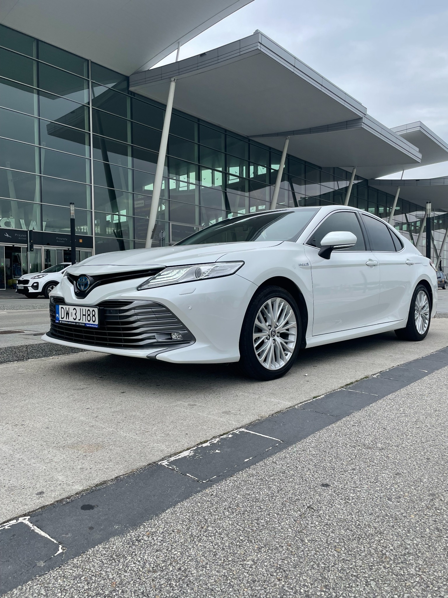 Biała Toyota Camry Hybrid zaparkowana przed nowoczesnym budynkiem lotniska z przeszkloną fasadą i charakterystycznymi daszkami.