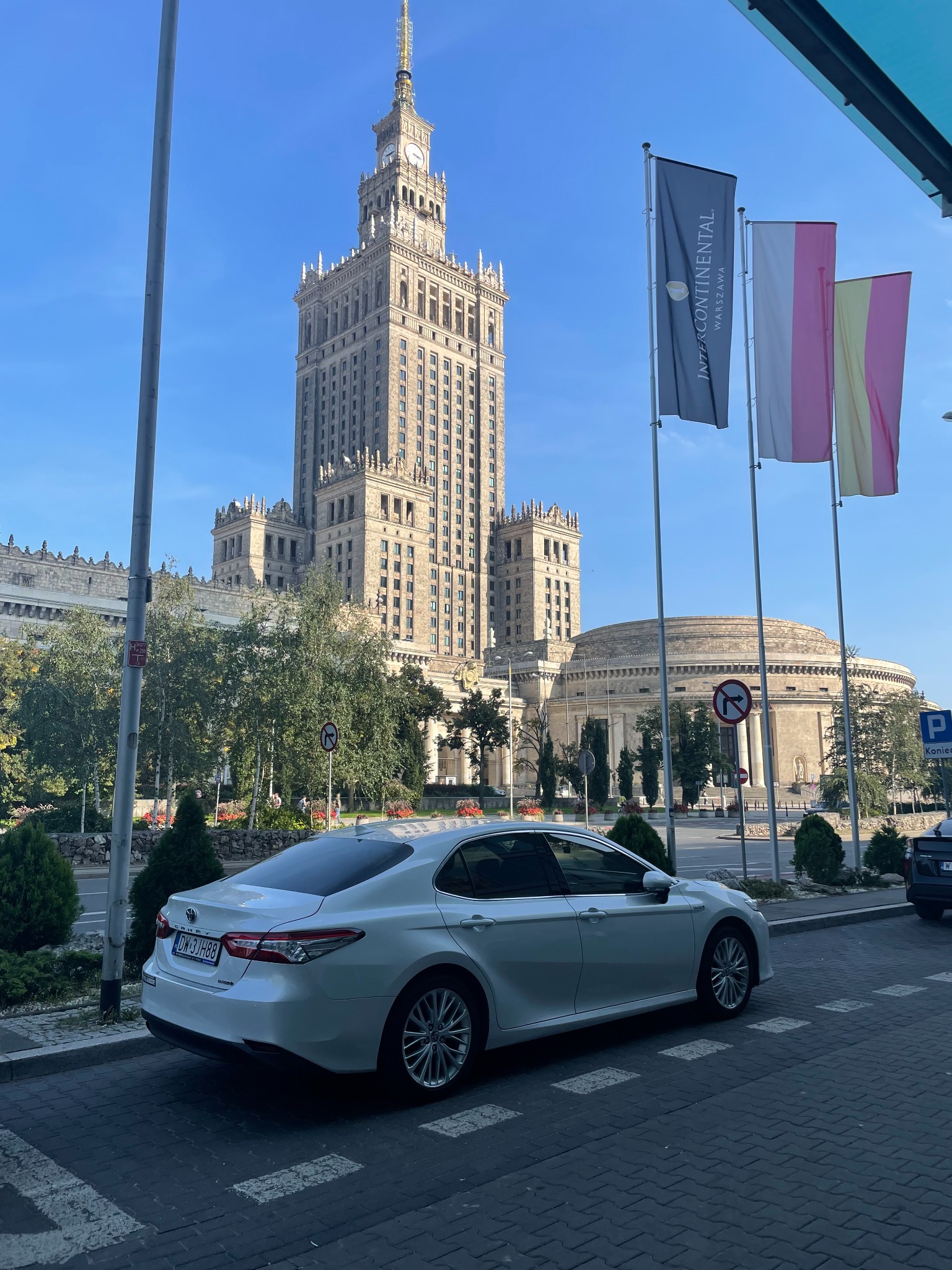Biała Toyota Camry zaparkowana na tle Pałacu Kultury i Nauki w Warszawie, z flagami InterContinental. Widoczne znaki drogowe i zieleń miejska.