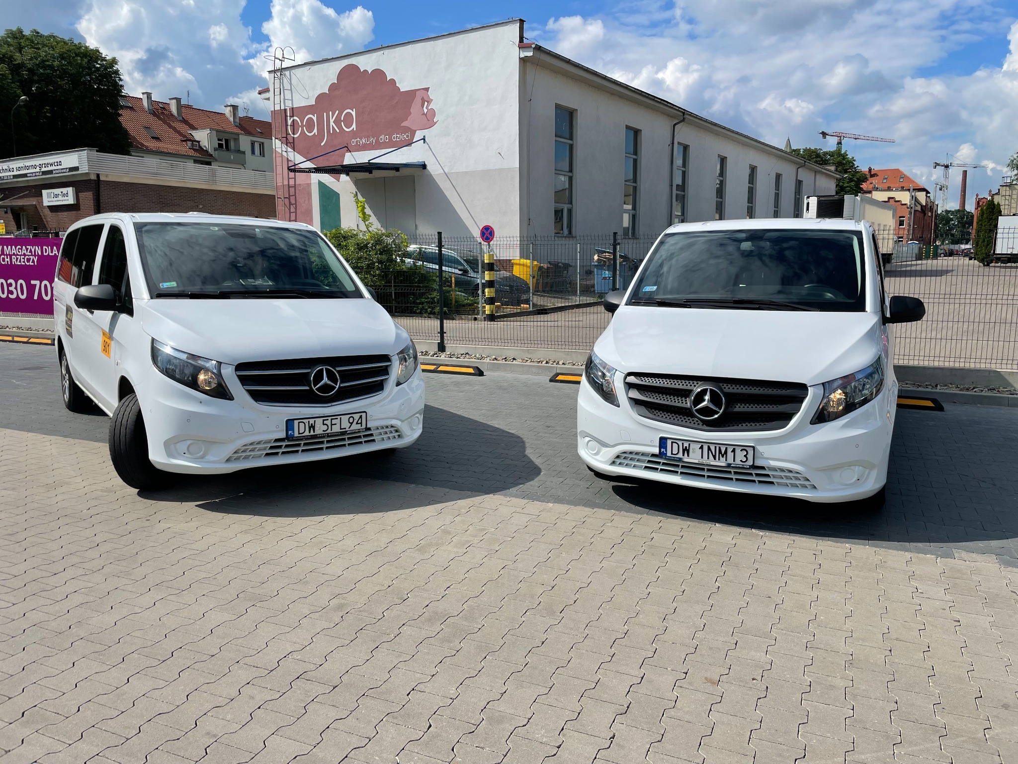 Dwa białe busy Mercedes Vito zaparkowane na brukowanym placu przed budynkiem z szyldem 'bajka artykuły dla dzieci'.