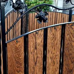 Drewniana brama z metalowymi zdobieniami w kształcie liści, prowadząca do ogrodu z widocznym iglakiem w tle.