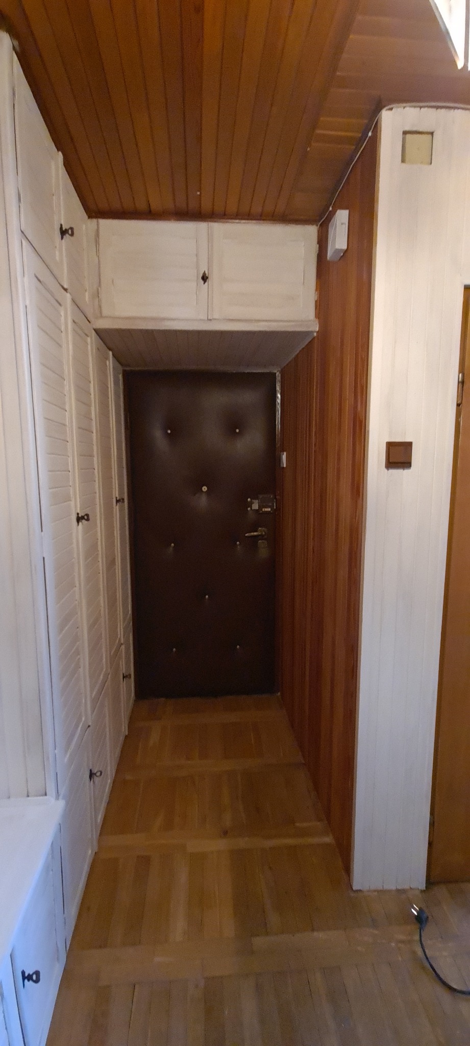 Wnętrze przedpokoju z drewnianą boazerią, podłogą i sufitem, zabudowaną szafą z białymi drzwiczkami oraz ciemnymi drzwiami wejściowymi z metalowymi okuciami.