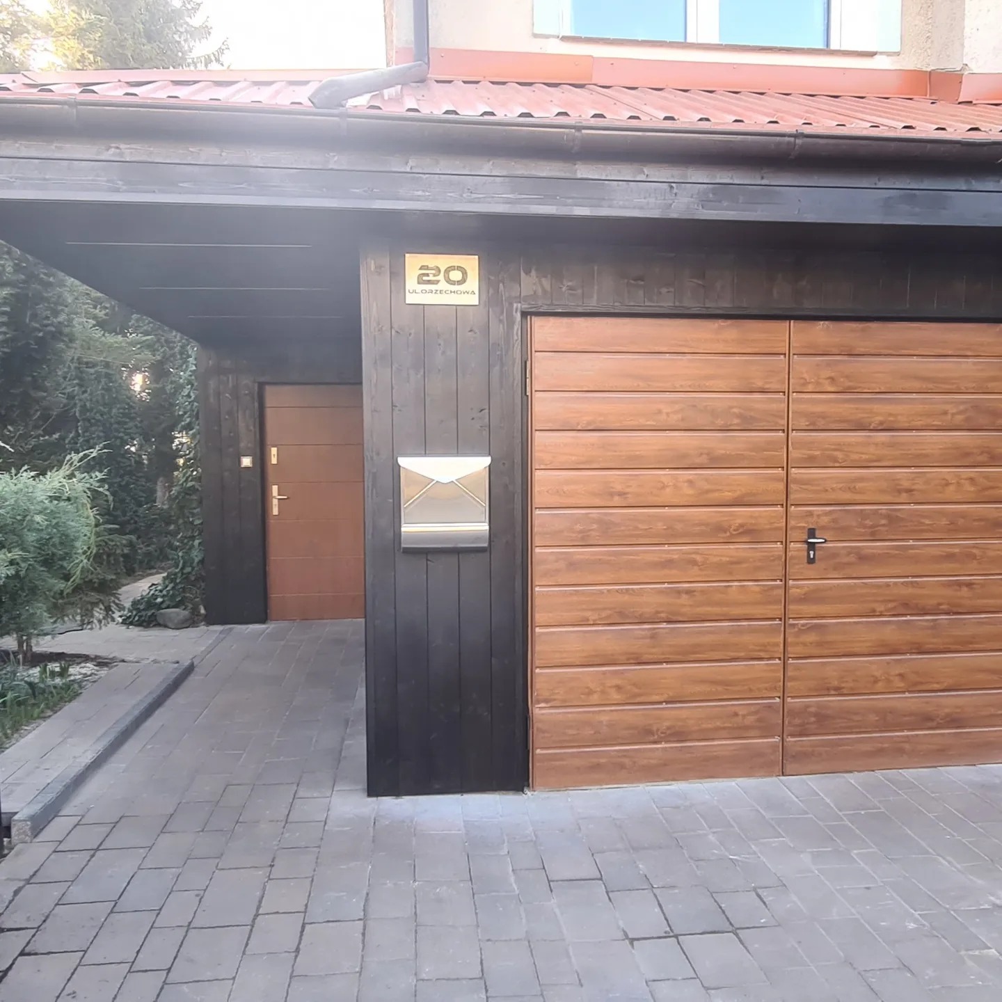 Drewniana brama garażowa w kolorze brązowym z poziomymi panelami, czarna drewniana konstrukcja wiaty garażowej z numerem domu na ścianie, szara kostka brukowa na podjeździe.