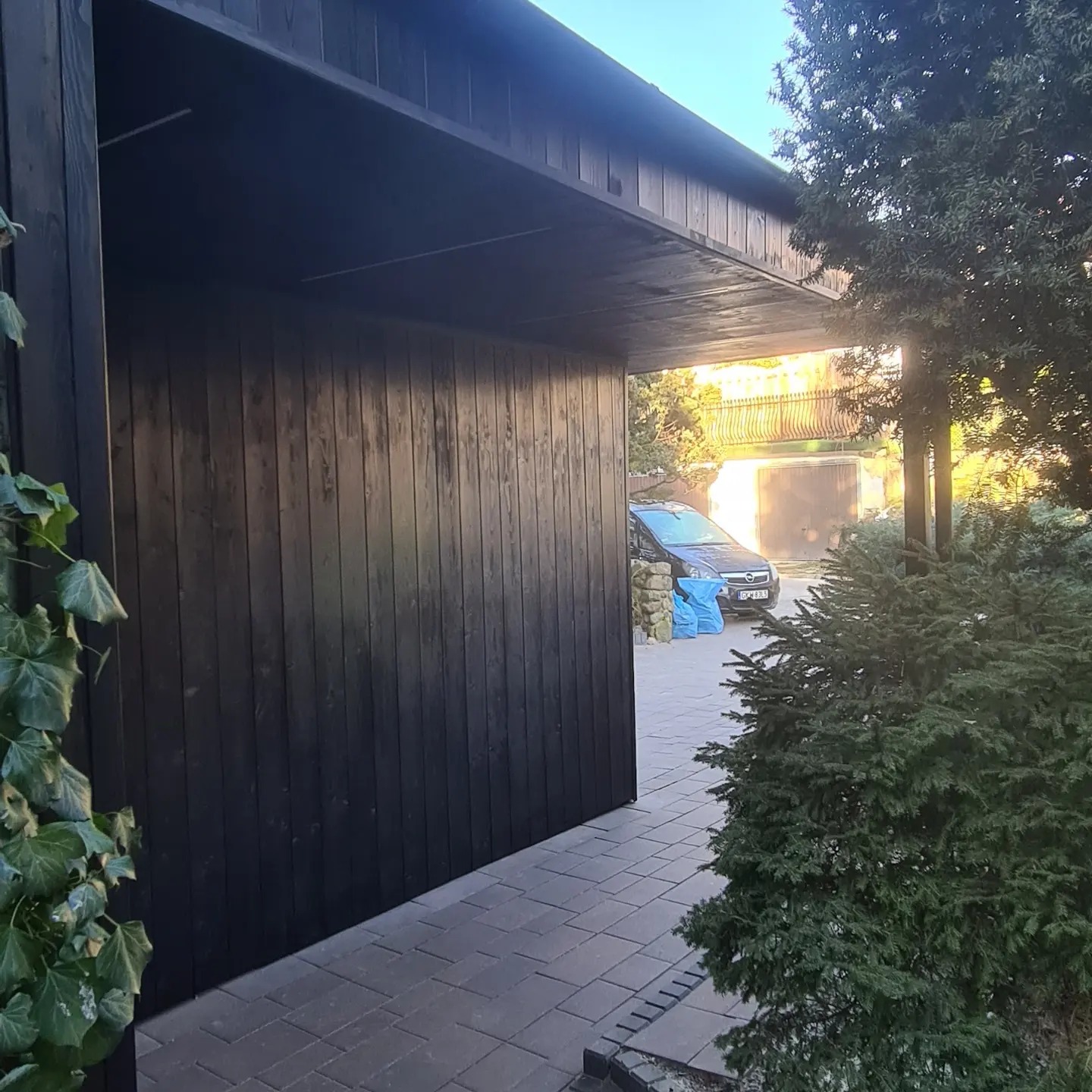 Nowoczesna, czarna, drewniana wiata garażowa z widocznym fragmentem podjazdu wyłożonego kostką brukową i samochodem w tle.