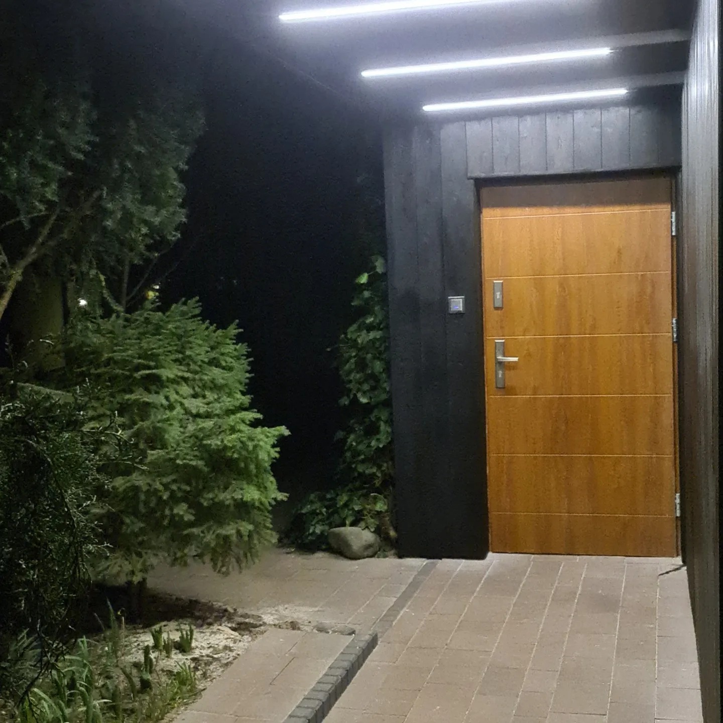 Wejście do domu z drewnianymi drzwiami oświetlonymi przez ledowe lampy, otoczone zielenią i wyłożone ceglaną kostką.