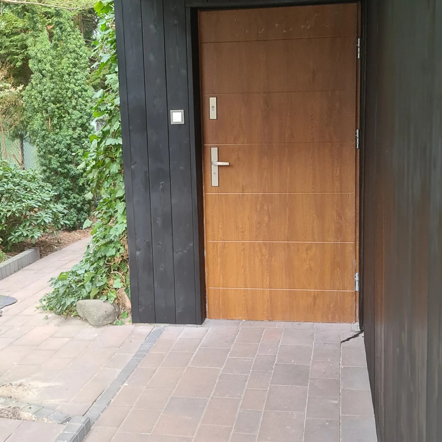 Nowoczesne, brązowe drzwi wejściowe z poziomymi panelami, osadzone w czarnej, drewnianej ramie, z widocznymi elementami okuć ze stali nierdzewnej, na tle kostki brukowej i zieleni.
