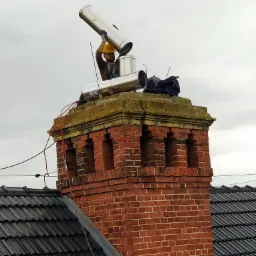 Pracownik w kasku montuje metalowy wkład kominowy na ceglanym kominie, widok z dołu na tle pochmurnego nieba.