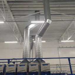 Instalacja wentylacyjna z rurami spiralnymi i szeregiem jednostek centralnych w hali przemysłowej, widok z dołu na biały sufit z oświetleniem LED.