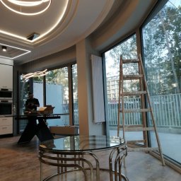 WELL DONE PAWEŁ MACIEJ JASIECKI - Jasne wnętrze z nowoczesnym oświetleniem sufitowym, drewnianą drabiną opartą o duże okno i pracującym mężczyzną w tle, częściowo wyposażone.