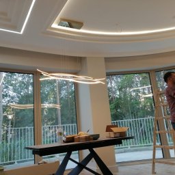 WELL DONE PAWEŁ MACIEJ JASIECKI - Pracownik na drabinie poprawia złote wykończenie sufitu podwieszanego z oświetleniem LED w przestronnym, jasnym pomieszczeniu z dużymi oknami.