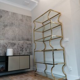 WELL DONE PAWEŁ MACIEJ JASIECKI - Elegancki regał z metalową ramą w kolorze złota i szklanymi półkami, ustawiony w nowoczesnym salonie z dekoracyjną ścianą i sztukaterią sufitową.