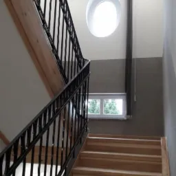 Klatka schodowa z drewnianymi schodami, czarną metalową balustradą i okrągłym oknem na półpiętrze, oświetlona lampami sufitowymi.