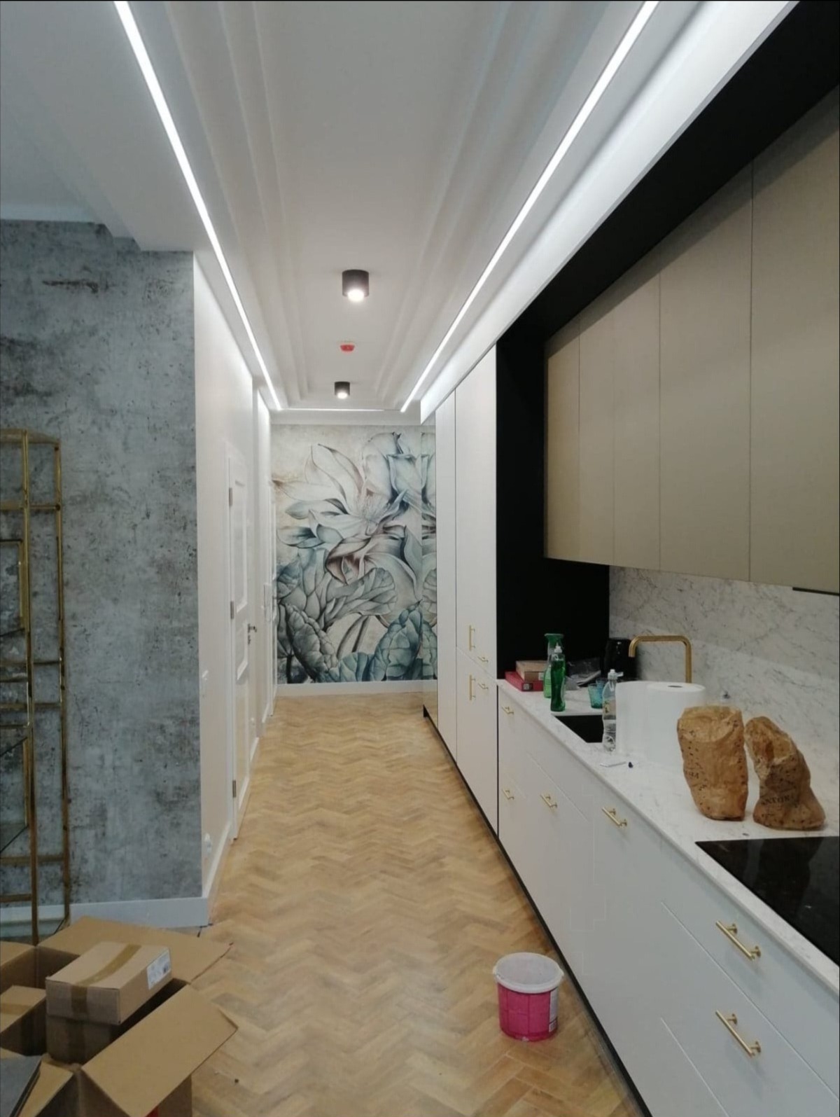 Nowoczesne wnętrze mieszkania z parkietem w jodełkę, oświetleniem LED podsufitowym, szafkami kuchennymi z białymi frontami i marmurowym blatem oraz betonową ścianą i fototapetą z motywem roślinnym...