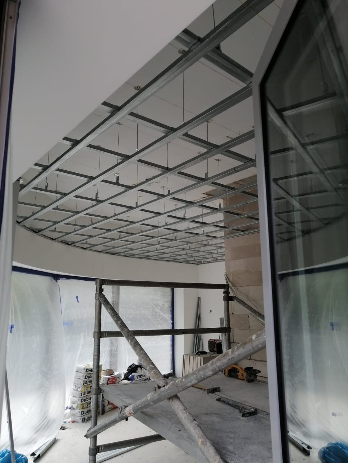 Montaż metalowej konstrukcji sufitu podwieszanego w pomieszczeniu z oknem, widoczne rusztowanie i worki z zaprawą.