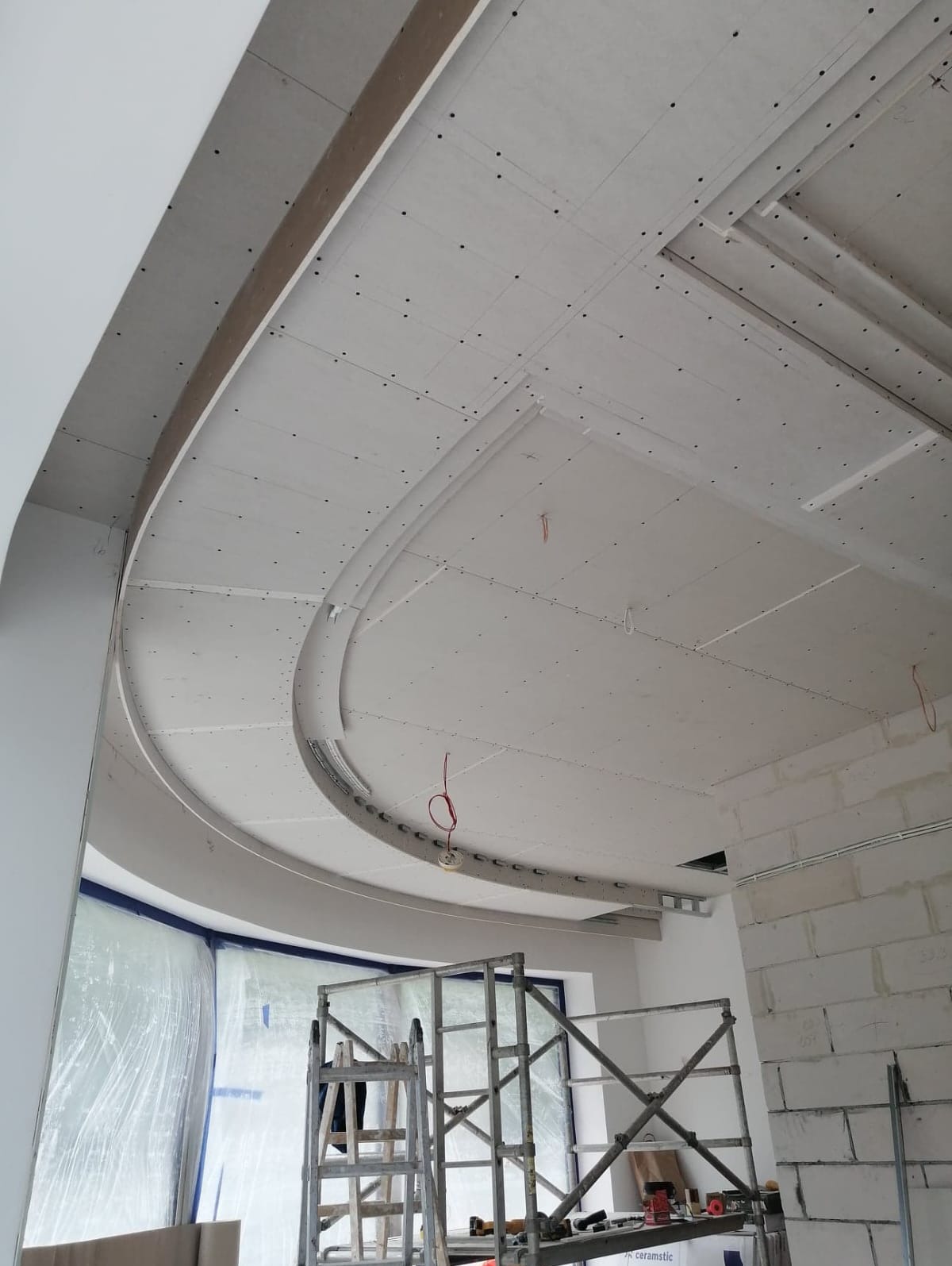 Niedokończony sufit z płyt kartonowo-gipsowych o zakrzywionym kształcie, widoczne metalowe profile, przewody elektryczne i rusztowanie w pomieszczeniu w trakcie budowy.