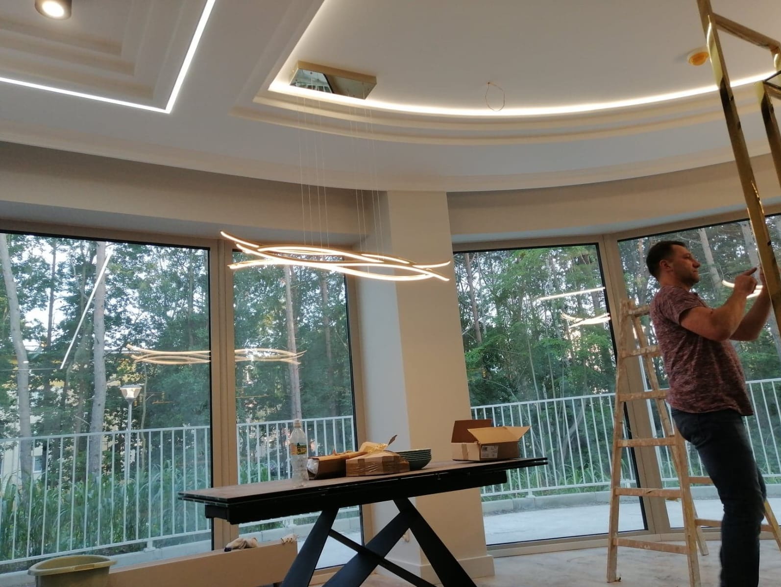 Pracownik na drabinie poprawia złote wykończenie sufitu podwieszanego z oświetleniem LED w przestronnym, jasnym pomieszczeniu z dużymi oknami.