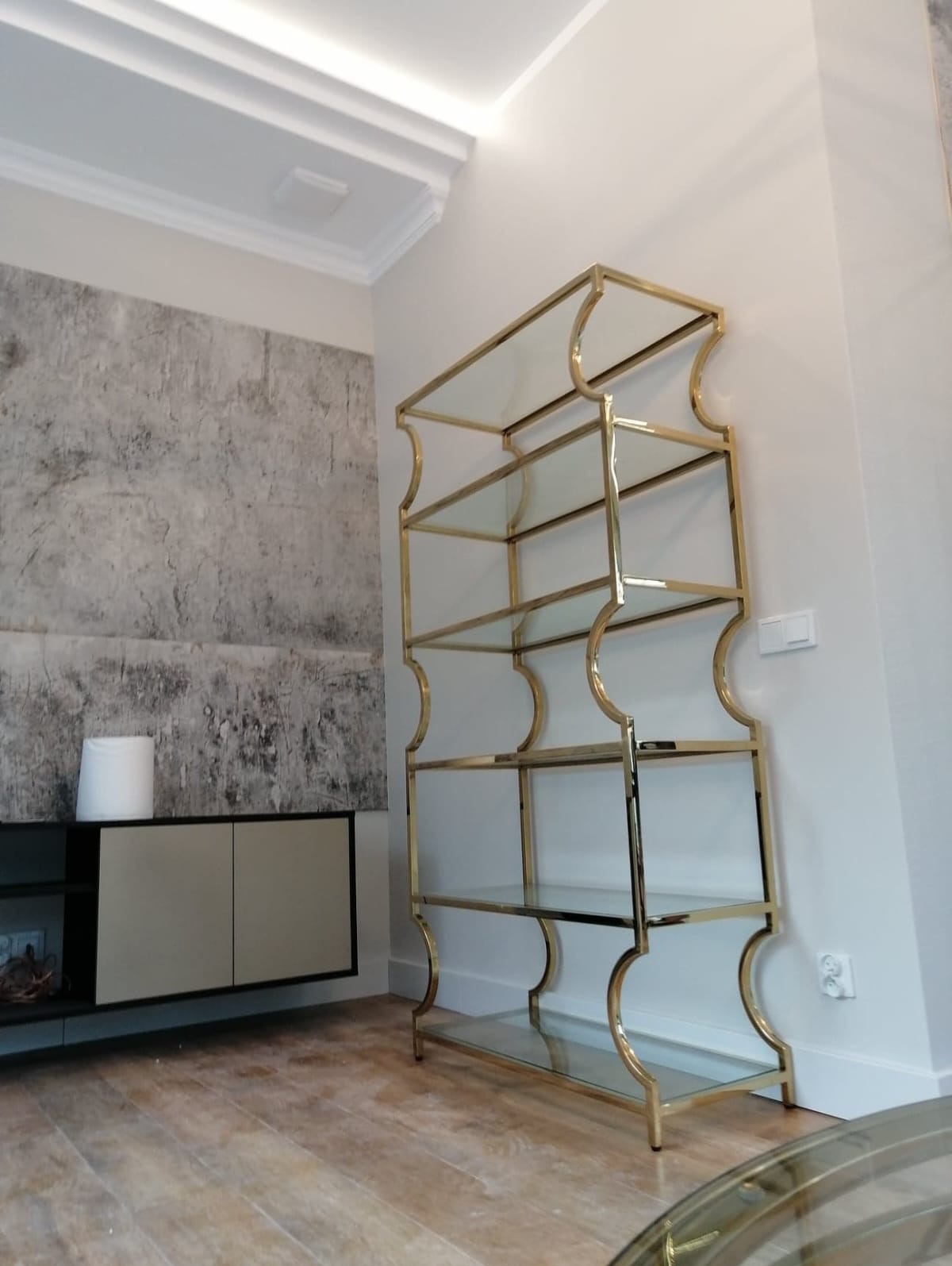 Elegancki regał z metalową ramą w kolorze złota i szklanymi półkami, ustawiony w nowoczesnym salonie z dekoracyjną ścianą i sztukaterią sufitową.