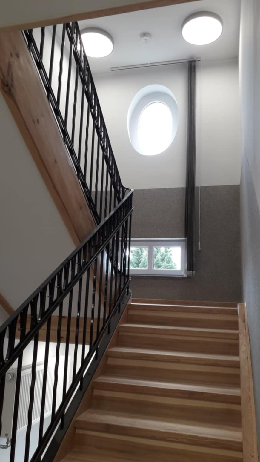 Klatka schodowa z drewnianymi schodami, czarną metalową balustradą i okrągłym oknem na półpiętrze, oświetlona lampami sufitowymi.