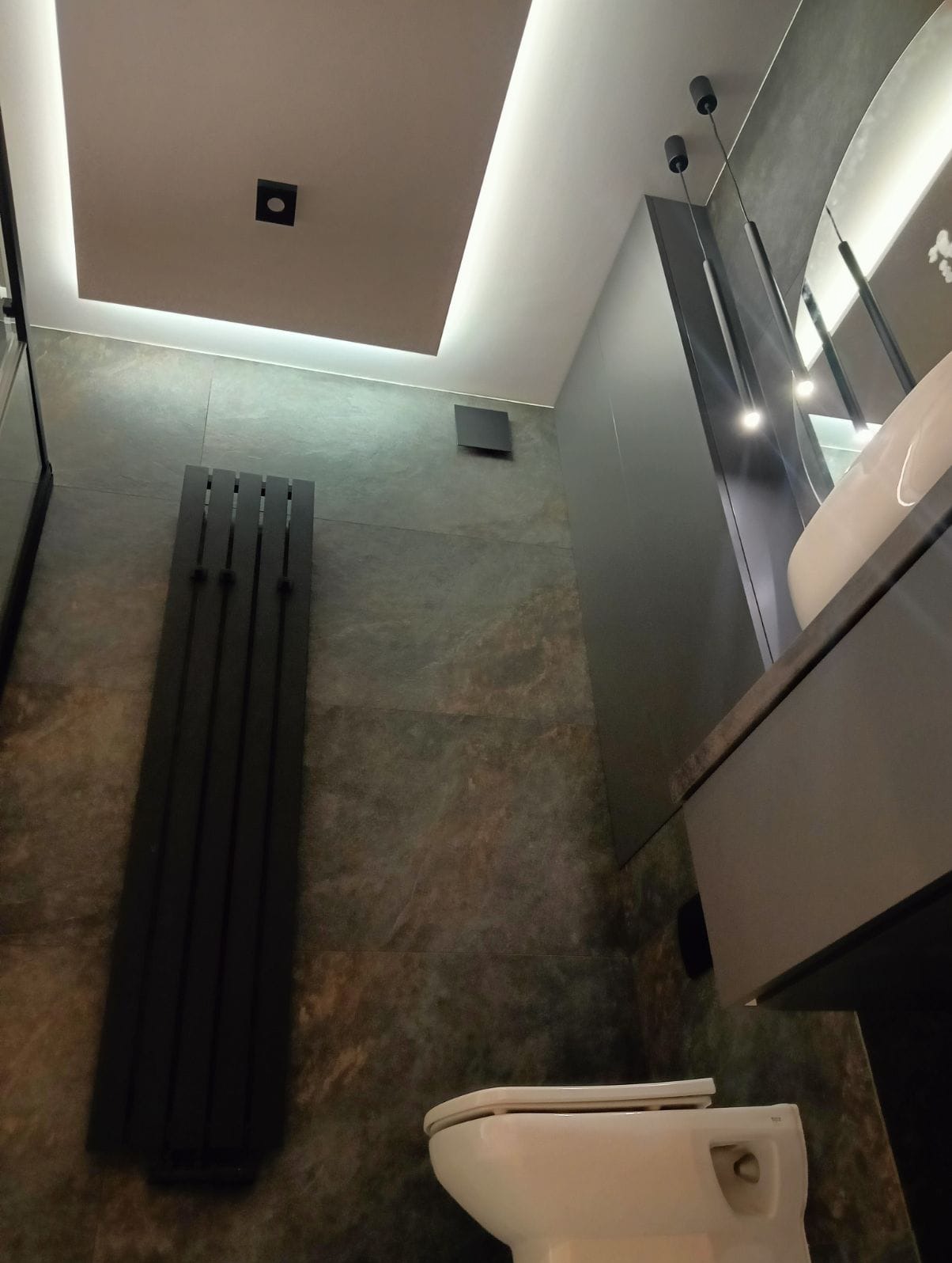 Nowoczesna łazienka z ciemnymi płytkami imitującymi kamień, podwieszana toaleta, grzejnik drabinkowy w kolorze czarnym i oświetlenie LED w suficie podwieszanym.
