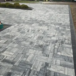 Ułożona, szara kostka brukowa w nieregularny wzór, obrzeża z ciemnej kostki. W tle ogród z krzewami i elementami architektury ogrodowej.