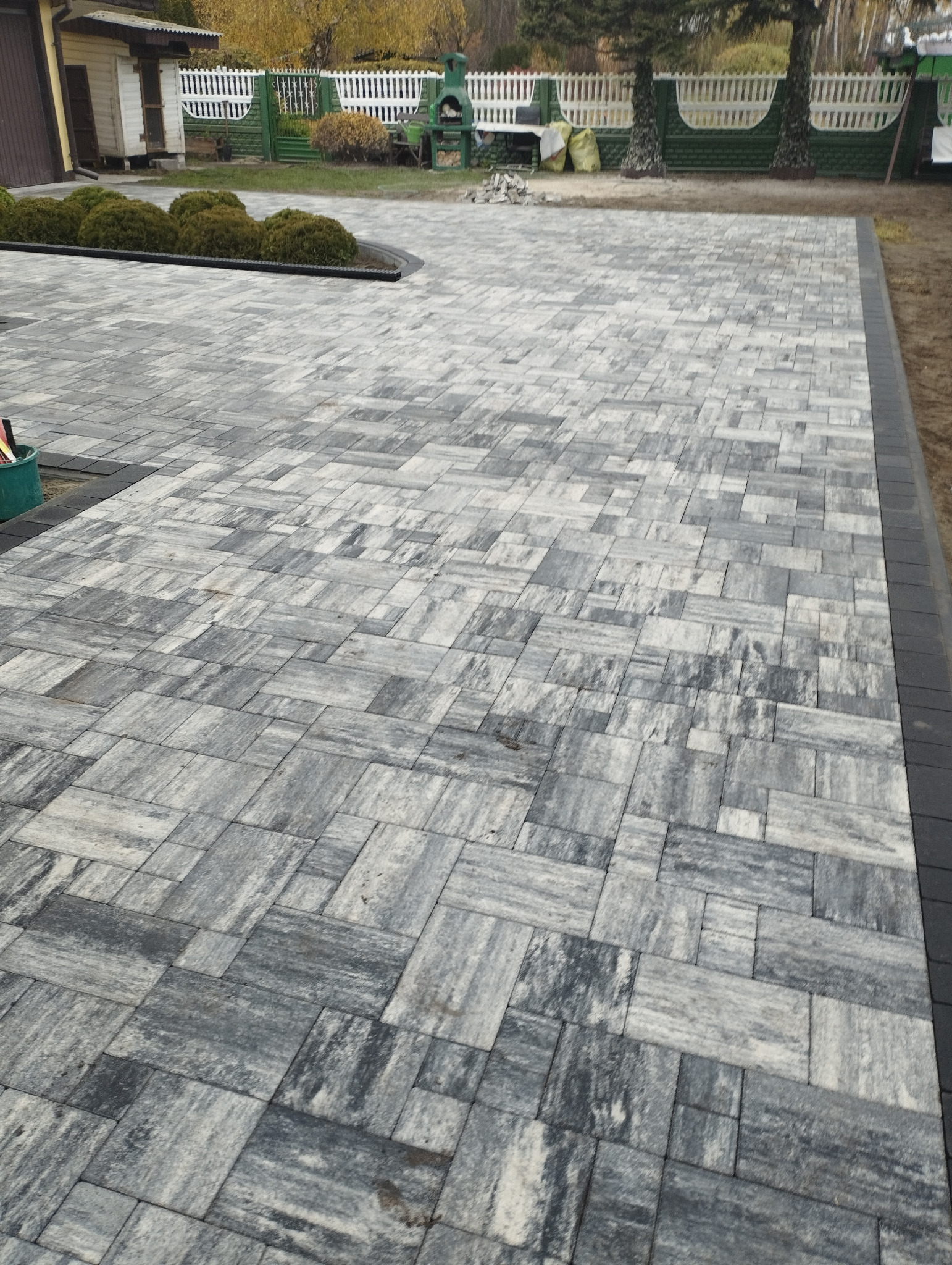 Ułożona, szara kostka brukowa w nieregularny wzór, obrzeża z ciemnej kostki. W tle ogród z krzewami i elementami architektury ogrodowej.