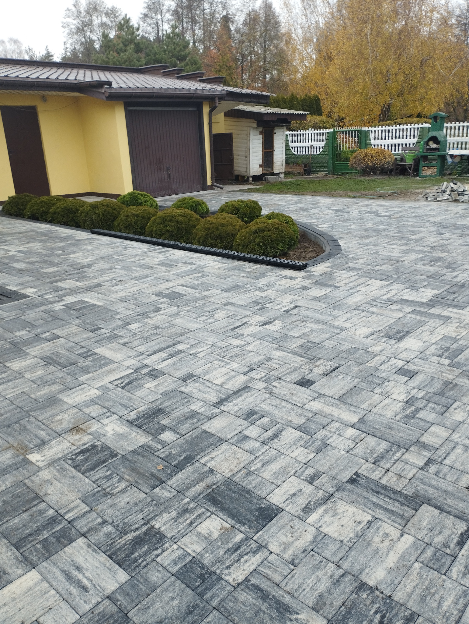Szara, ułożona kostka na posesji z obrzeżem i rabatą. W tle budynek gospodarczy i drzewa jesienią. Widok z góry na gotową nawierzchnię.