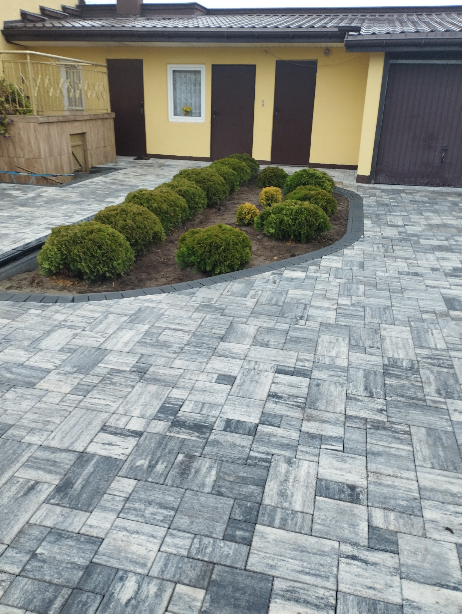 Szara kostka brukowa ułożona wzorem jodełki przed budynkiem z żółtą elewacją i szpalerem kulistych krzewów. Estetyczne wykończenie posesji.