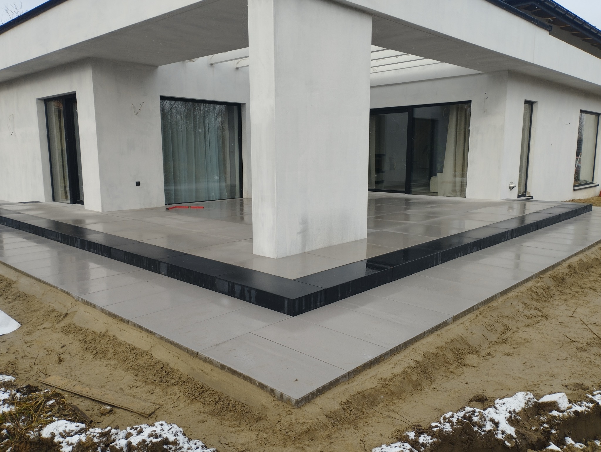 Nowoczesny taras z jasnoszarej kostki, obrzeża z ciemnej kostki, minimalistyczna architektura budynku w tle. Widoczny fragment gruntu z resztkami śniegu.