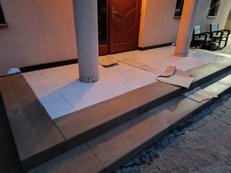 Wykończone schody wejściowe z płytkami w odcieniach brązu i beżu, z widocznymi kolumnami i fragmentami zabezpieczeń na powierzchni.