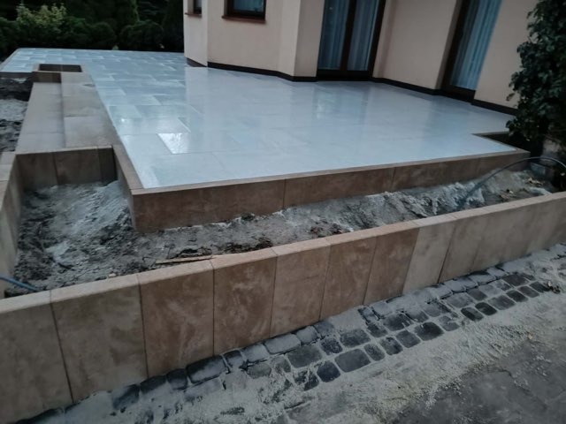 Jasne płytki tarasowe przylegają do budynku. Obok, obrzeże z płytek w odcieniach beżu, oddziela taras od ziemi. Widoczne fragmenty kostki brukowej.