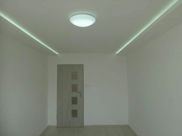 Pusty pokój z białymi ścianami, panelami LED w suficie i szarymi drzwiami z czterema szybkami.