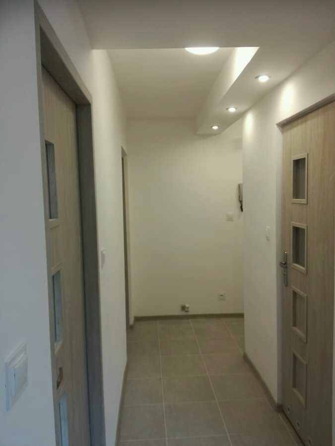 Wnętrze korytarza po remoncie z dwiema drewnianymi ościeżnicami, szarą podłogą z płytek i białymi ścianami z oświetleniem sufitowym.