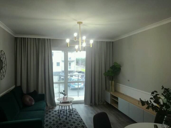 Jasne wnętrze pokoju dziennego z szarymi ścianami, zasłonami i podłogą, ciemnozielona sofa z okrągłą poduszką, złoty żyrandol sufitowy, stoliki kawowe, szafka RTV z roślinami doniczkowymi