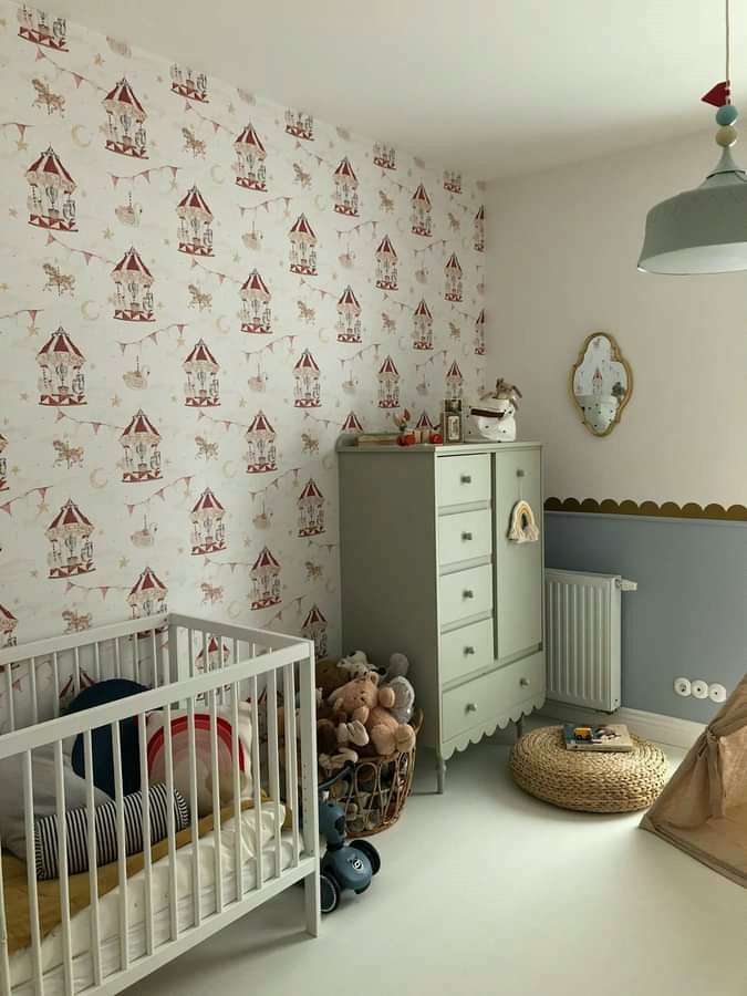 Pokój dziecięcy z tapetą w motywy karuzeli, białe łóżeczko z kolorowymi poduszkami, szara komoda z dekoracjami, pleciony puf i fragment tipi.