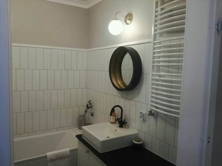 Biała łazienka z pionowo ułożonymi płytkami, okrągłym lustrem w czarnej ramie z złotym wnętrzem, grzejnikiem drabinkowym i umywalką na czarnym blacie.