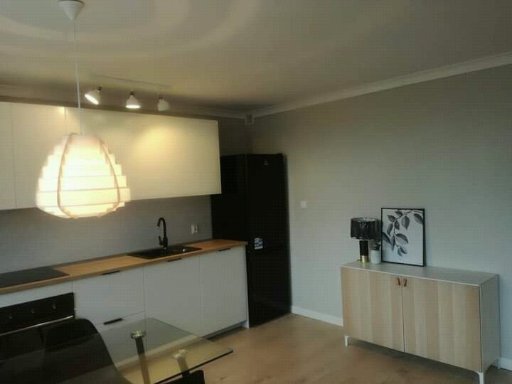 Nowoczesna kuchnia z białymi szafkami, drewnianym blatem i czarną lodówką, oświetlona designerską lampą wiszącą i reflektorami