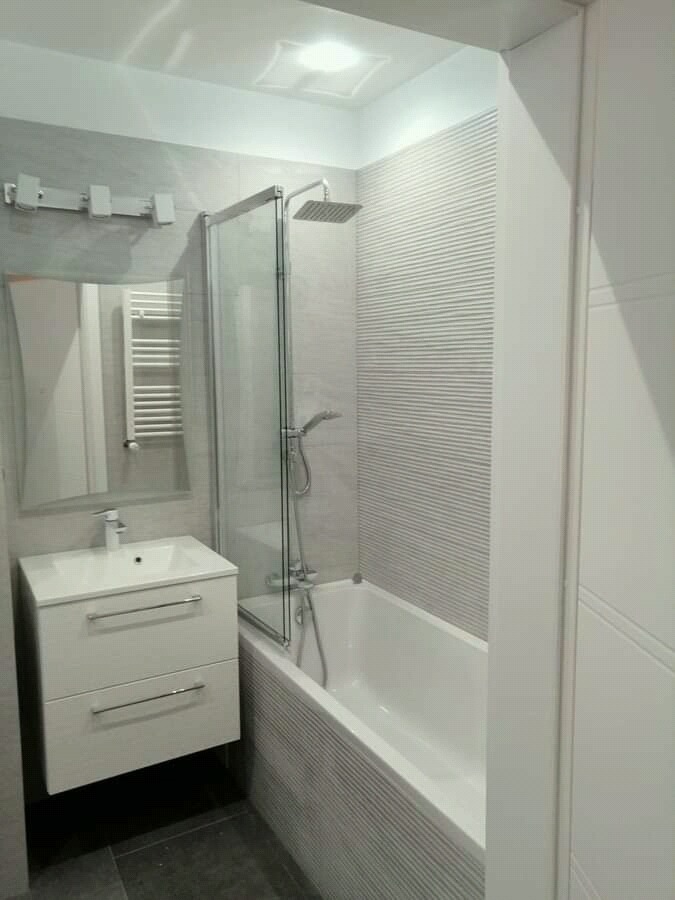 Wykończona łazienka w odcieniach szarości z białą armaturą: wanna z szklanym parawanem, umywalka z szafką i grzejnik drabinkowy.