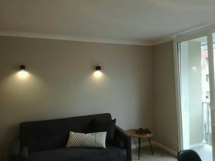 Pokój dzienny po odświeżeniu: szare ściany, czarna sofa z poduszką w geometryczne wzory, dwie nowoczesne lampy ścienne, okno balkonowe z widokiem na budynek.