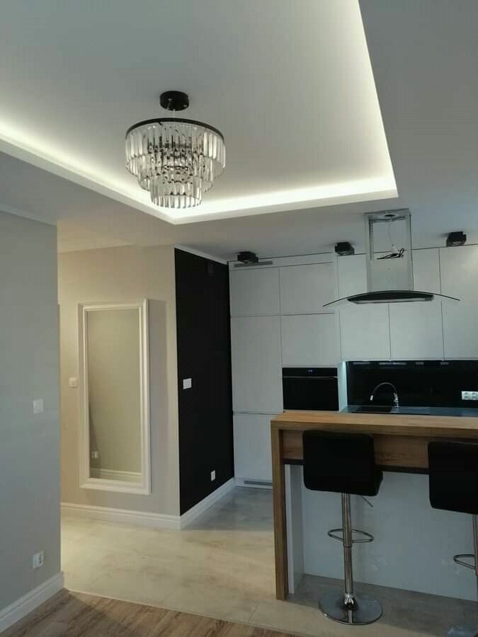 Nowoczesna kuchnia z białymi szafkami, czarnym panelem nad blatem, drewnianym barkiem z hokerami i designerskim żyrandolem kryształowym podświetlonym ledami w suficie podwieszanym.