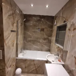 Wykończona łazienka z wanną, umywalką i marmurowymi płytkami w odcieniach beżu i brązu, oświetlona punktowymi lampami sufitowymi i dodatkową lampą nad lustrem.